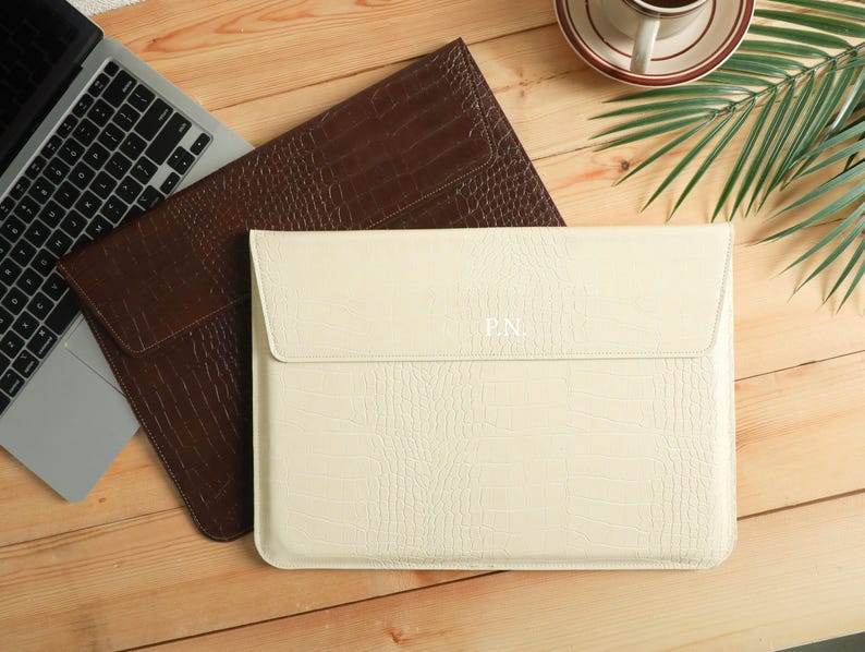 Op de afbeelding: Twee laptoptassen met een krokodillenleertextuur. E&eacute;n is cr&egrave;mekleurig met de initialen "P.N." in reli&euml;f op de voorkant. De andere is bruin. Een laptop en een kopje koffie zijn ook zichtbaar op het houten oppervlak.