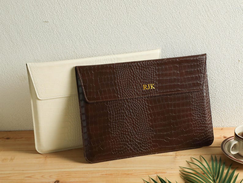Op de afbeelding: Twee laptoptassen. E&eacute;n is cr&egrave;mekleurig, de andere is bruin met een krokodillenstructuur. De bruine tas heeft de initialen "RJK" in gouden letters. Beide tassen liggen op een houten oppervlak.