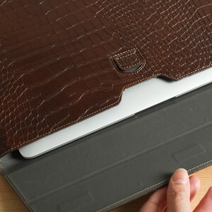 Op de afbeelding: Een bruine, getextureerde leren laptoptas met een krokodillenleerpatroon. De tas is open en onthult een zilveren laptop binnenin. De binnenkant is een donkergrijze stof. De tas ligt op een lichtbruin houten oppervlak.