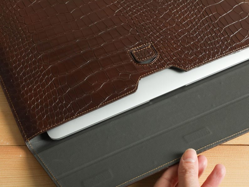 Op de afbeelding: Een bruine, getextureerde leren laptoptas met een krokodillenleerpatroon. De tas is open en onthult een zilveren laptop binnenin. De binnenkant is een donkergrijze stof. De tas ligt op een lichtbruin houten oppervlak.