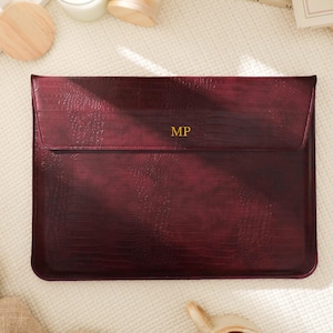 Könnte beinhalten: Eine weinrote Laptop-Hülle aus Leder mit einem strukturierten, geprägten Muster. Der Klappenverschluss trägt die goldenen Initialen "MP". Die Hülle ist rechteckig und zum Schutz eines Laptops konzipiert. Die Gesamtästhetik ist elegant und professionell.