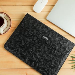 Op de afbeelding: Zwarte laptoptas met een reli&euml;f bloemenpatroon, het woord "KAMI" is zichtbaar. Een witte computermuis, een laptop en een kopje thee op een schotel liggen ook op een houten oppervlak.
