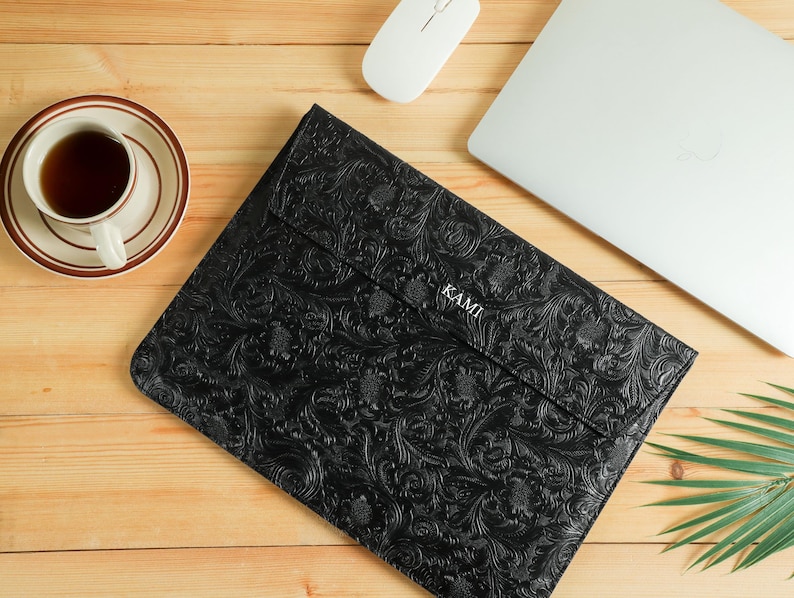 Op de afbeelding: Zwarte laptoptas met een reli&euml;f bloemenpatroon, het woord "KAMI" is zichtbaar. Een witte computermuis, een laptop en een kopje thee op een schotel liggen ook op een houten oppervlak.