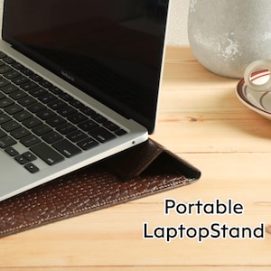 Op de afbeelding: Een zilveren laptop rust op een bruine leren laptopstandaard met een textuur. De standaard staat op een licht houten oppervlak. Een kopje thee staat rechts op een schotel. Tekst op de standaard luidt "Portable LaptopStand."