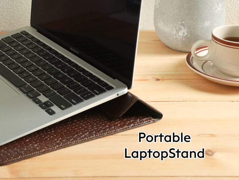 Op de afbeelding: Een zilveren laptop rust op een bruine leren laptopstandaard met een textuur. De standaard staat op een licht houten oppervlak. Een kopje thee staat rechts op een schotel. Tekst op de standaard luidt "Portable LaptopStand."