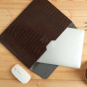 Op de afbeelding: Een bruine leren laptoptas met krokodillenleertextuur, met een laptop erin. Een witte draadloze muis en een kopje koffie staan op een houten oppervlak. De tas is ontworpen om een laptop te beschermen.