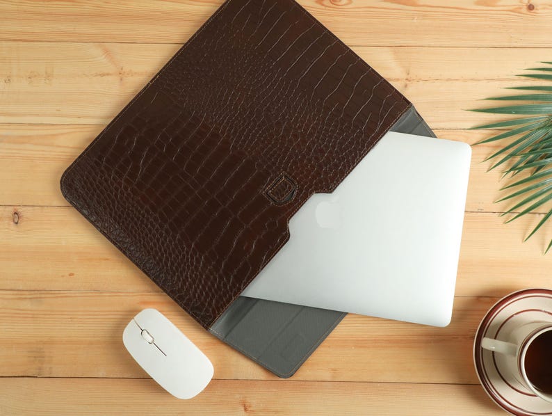 Op de afbeelding: Een bruine leren laptoptas met krokodillenleertextuur, met een laptop erin. Een witte draadloze muis en een kopje koffie staan op een houten oppervlak. De tas is ontworpen om een laptop te beschermen.