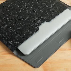 Op de afbeelding: Zwarte leren laptoptas met een reli&euml;f bloemenpatroon. Een zilveren laptop is gedeeltelijk zichtbaar in de hoes, die op een grijze tablethoes rust. Een witte computermuis bevindt zich aan de rechterkant.