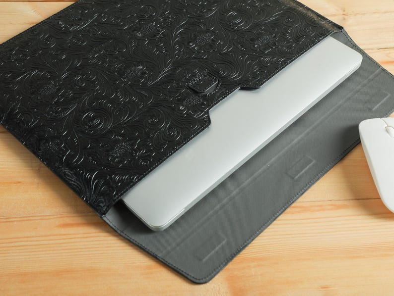 Op de afbeelding: Zwarte leren laptoptas met een reli&euml;f bloemenpatroon. Een zilveren laptop is gedeeltelijk zichtbaar in de hoes, die op een grijze tablethoes rust. Een witte computermuis bevindt zich aan de rechterkant.