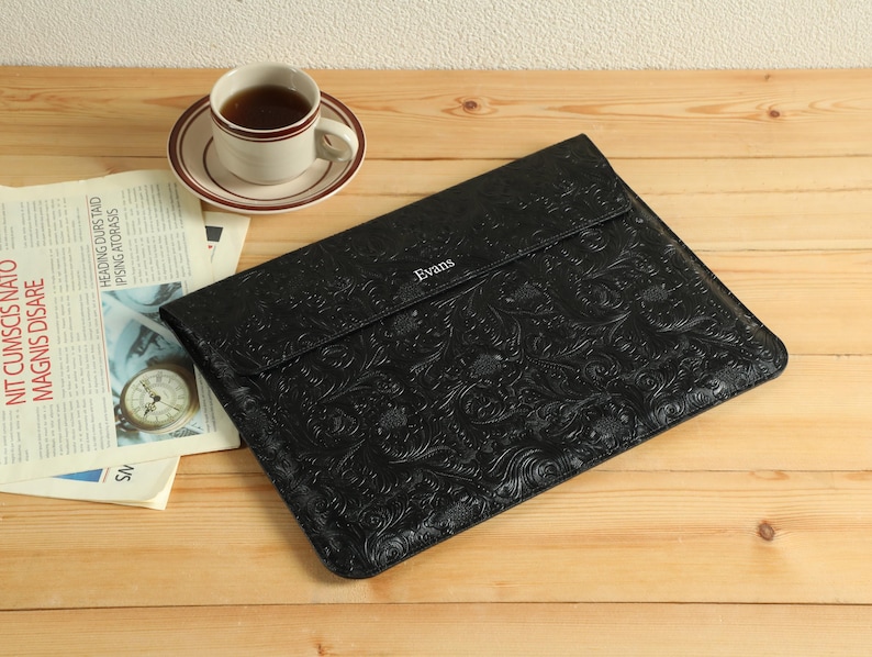 Op de afbeelding: Een zwarte leren laptoptas met een reli&euml;f bloemmotief, het woord "Evans" is in de flap ge&euml;mbosseerd. Een kopje thee op een schotel en een krant liggen op een houten oppervlak.