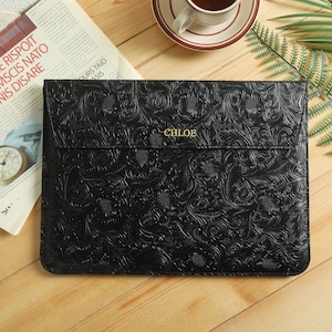 Op de afbeelding: Zwarte laptoptas met een reli&euml;f bloemenpatroon en de naam "CHLOE" in gouden letters. De gesloten tas ligt op een houten oppervlak, naast een krant en een kopje koffie.
