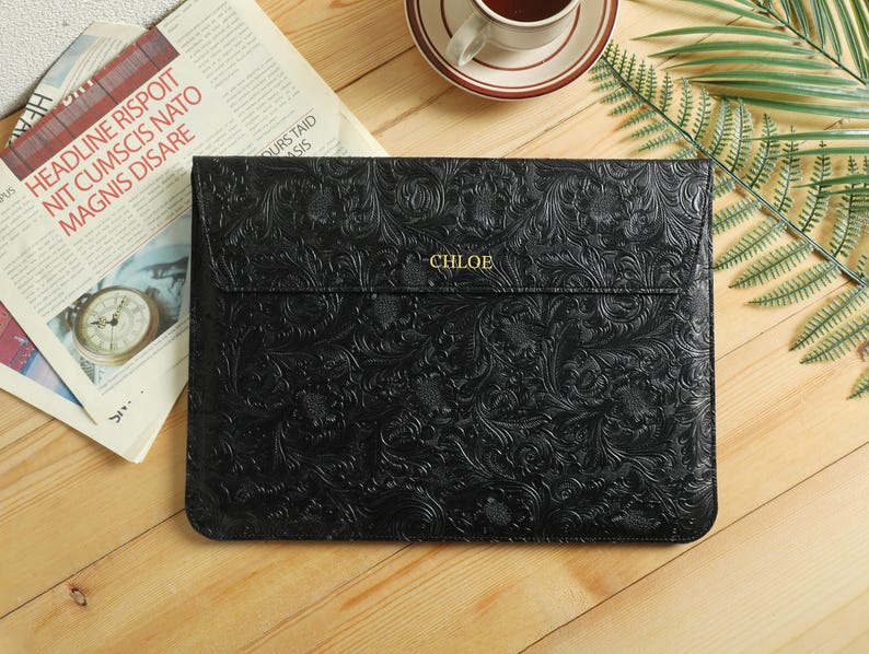 Op de afbeelding: Zwarte laptoptas met een reli&euml;f bloemenpatroon en de naam "CHLOE" in gouden letters. De gesloten tas ligt op een houten oppervlak, naast een krant en een kopje koffie.
