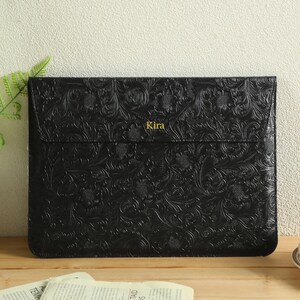 Op de afbeelding: Zwarte leren laptoptas met een reli&euml;f bloemenpatroon. Het woord "Kira" is in goud op de voorkant gedrukt. De tas is rechthoekig en heeft een klepsluiting. Een kopje koffie en een plant staan op de achtergrond.