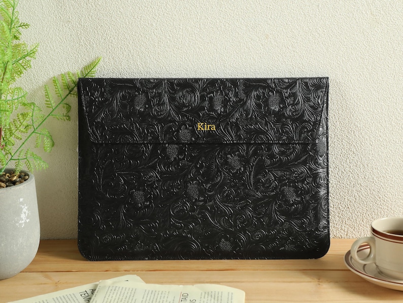 Op de afbeelding: Zwarte leren laptoptas met een reli&euml;f bloemenpatroon. Het woord "Kira" is in goud op de voorkant gedrukt. De tas is rechthoekig en heeft een klepsluiting. Een kopje koffie en een plant staan op de achtergrond.