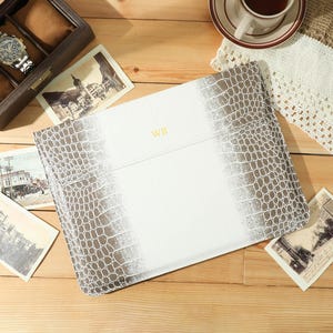 May include: A white and gray faux-crocodile leather laptop sleeve with the initials "WR" in gold. The sleeve is on a wooden surface with vintage postcards, a watch in a wooden box, and a cup of coffee.