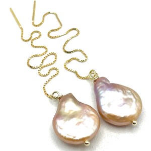May include: A pair of gold-toned drop earrings featuring large, irregularly shaped, pinkish-cream pearls. The earrings have a delicate chain design that extends upwards, creating a flowing, elegant look. The pearls have a lustrous, iridescent quality.