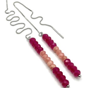 Puede incluir: Pendientes colgantes con un degradado de cuentas rosas y melocotón. Las cuentas están ensartadas en una cadena plateada que se curva y termina con un poste recto. Los pendientes están sobre un fondo blanco.