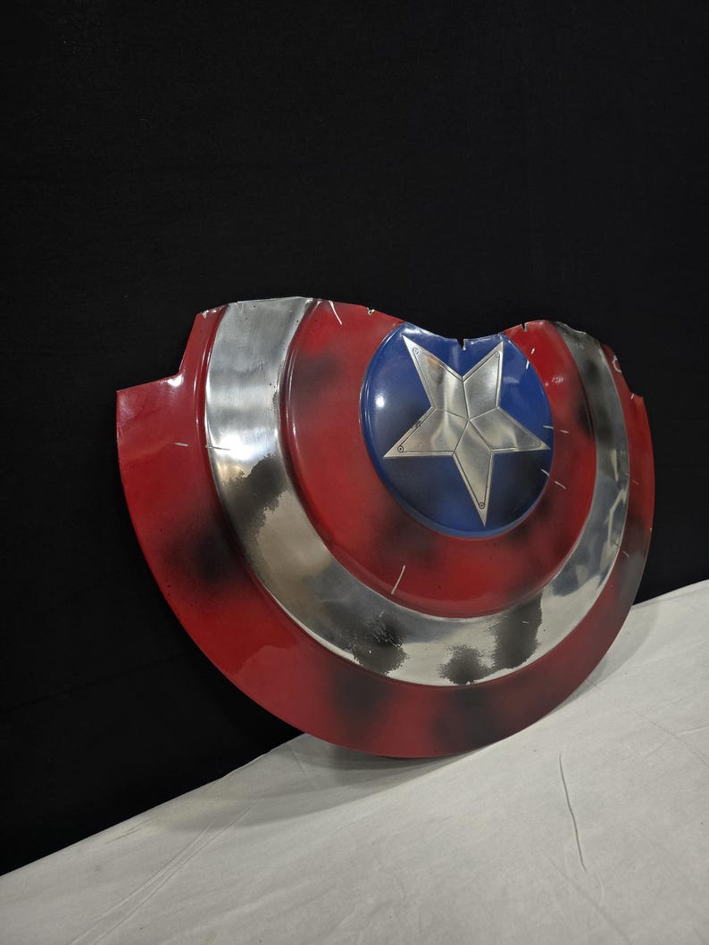 May include: A red, white, and blue shield with a silver star in the center. The shield has a circular design with a silver band and a blue circle in the middle. The shield has a distressed look.