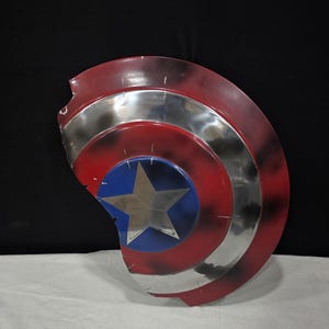 May include: A distressed Captain America shield, featuring a red, white, and blue color scheme. The shield has a silver metallic band and a blue center with a silver star. The shield shows signs of wear and tear.