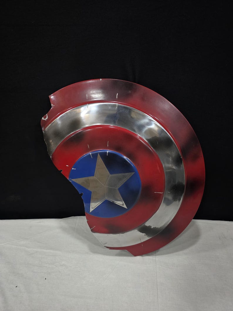 May include: A distressed Captain America shield, featuring a red, white, and blue color scheme. The shield has a silver metallic band and a blue center with a silver star. The shield shows signs of wear and tear.