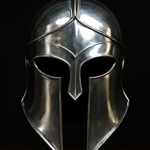 Puede incluir: Un casco espartano plateado, con una prominente cresta frontal, aberturas para los ojos y una pluma vertical. La superficie del casco refleja la luz, creando reflejos y sombras sobre un fondo negro. Objeto histórico.