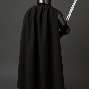 May include: A person in medieval armor, holding a long sword. The figure is wearing a black cloak, a metal helmet, and gauntlets. The armor has a metallic finish, and the sword is silver. The background is a solid gray.