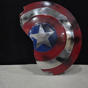 May include: A distressed Captain America shield, featuring a red, white, and blue color scheme. The shield has a silver star in the center, surrounded by a blue circle. The outer edges are red with silver stripes, and the shield has a damaged appearance.
