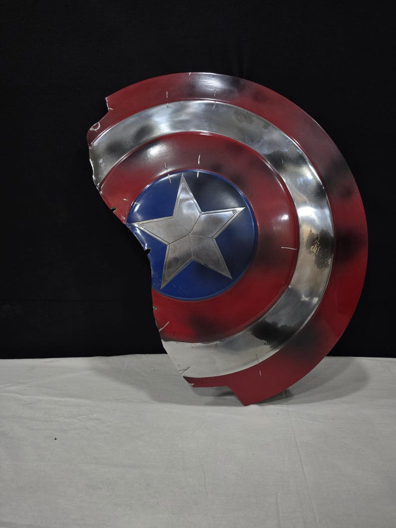 May include: A distressed Captain America shield, featuring a red, white, and blue color scheme. The shield has a silver star in the center, surrounded by a blue circle. The outer edges are red with silver stripes, and the shield has a damaged appearance.