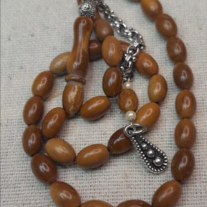 May include: A prayer bead necklace with oval-shaped, light brown beads. The beads are strung together with a silver chain and a decorative silver pendant. The tassel is made of wood.