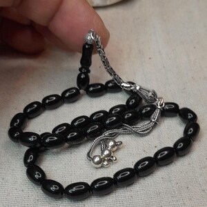 Peut inclure: Chapelet noir avec des perles ovales et des détails argentés. Les perles sont enfilées sur une chaîne et un pompon argentés. Les perles sont tenues par une main.