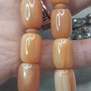 Op de afbeelding: Close-up van een streng lichtoranje, tonvormige kralen. De kralen hebben een glanzende afwerking en zijn aan elkaar geregen. De achtergrond is een wazig beeld van een hand en andere sieraden.