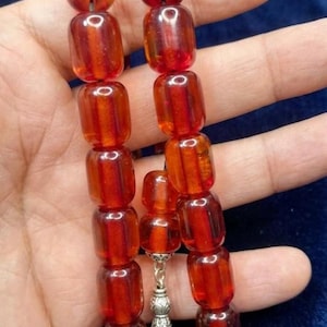 May include: A set of amber-coloured prayer beads, each bead is a rounded rectangle. The beads are strung together with a silver-coloured tassel and decorative piece. The beads are a translucent, reddish-brown colour.