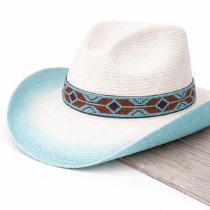 Peut inclure: Un chapeau de cowboy blanc avec un bord bleu clair en dégradé. Le chapeau est orné d'un bandeau décoratif avec un motif géométrique marron, turquoise et violet. Le chapeau est en matière tissée.