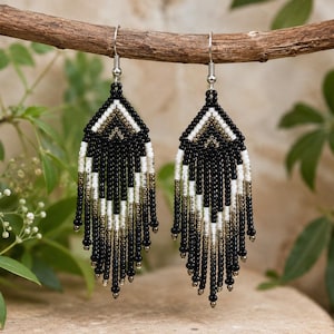 Close-up of handmade black, white, and metallic seed bead fringe earrings hanging from a rustic wooden branch in natural light.