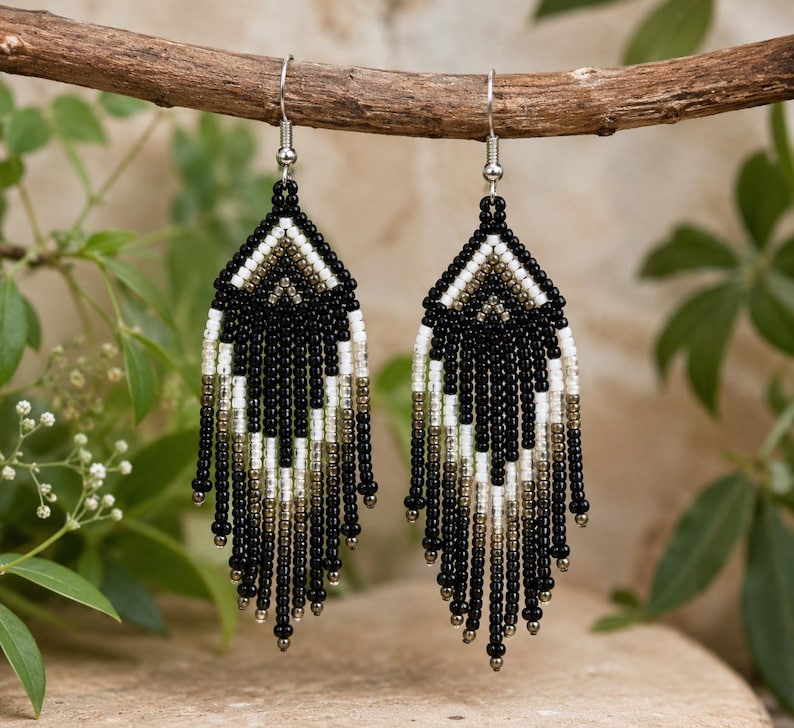 Close-up of handmade black, white, and metallic seed bead fringe earrings hanging from a rustic wooden branch in natural light.