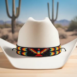 Może przedstawiać: Biały kapelusz kowbojski z paciorkową opaską z kolorowym geometrycznym wzorem. Opaska zawiera czerwone, żółte i niebieskie akcenty na czarnym tle. Kapelusz jest osadzony na tle pustynnego krajobrazu.