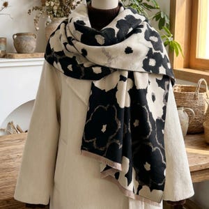 May include: A cream-colored scarf with a bold black floral pattern is draped over a cream-colored coat. The scarf's design features large, stylized flowers. The coat has a classic, tailored design. The scarf is made of a soft material and has a fringed edge.