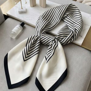May include: A square scarf featuring a cream-colored base with black stripes. The pattern consists of thin parallel lines that transition into a chevron design near the corners, which are finished with a solid black border along the edges.
