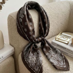 May include: A square scarf in a deep chocolate brown color featuring a white paisley and floral pattern. The scarf is tied in a knot, displaying a repeating motif across the fabric with a decorative border design along the edges of the material.