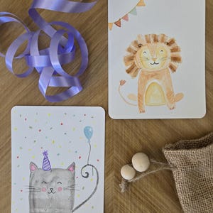 Peut inclure: Deux cartes de vœux illustrées aux coins arrondis. L'une représente un lion souriant avec une bannière, l'autre un chat avec un chapeau de fête et un ballon. Un ruban violet, des perles en bois et un sac en toile de jute sont également présents.