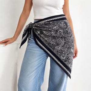 May include: A black and white paisley print scarf tied around the waist over light blue denim jeans and a white tank top. The scarf features a detailed intricate pattern with a solid black border and a white geometric trim along the edges.