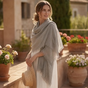 May include: A light gray, rectangular scarf with a textured, woven appearance and frayed edges, draped over the shoulders. The scarf is made of a lightweight fabric and is worn over a simple white garment, paired with a straw hat held in one hand.