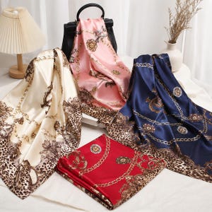 May include: Four square satin scarves in cream, pink, navy blue, and red, each featuring a repeating pattern of gold-toned chains, circular medallions, and brown leopard print borders. The scarves are displayed draped and folded together.