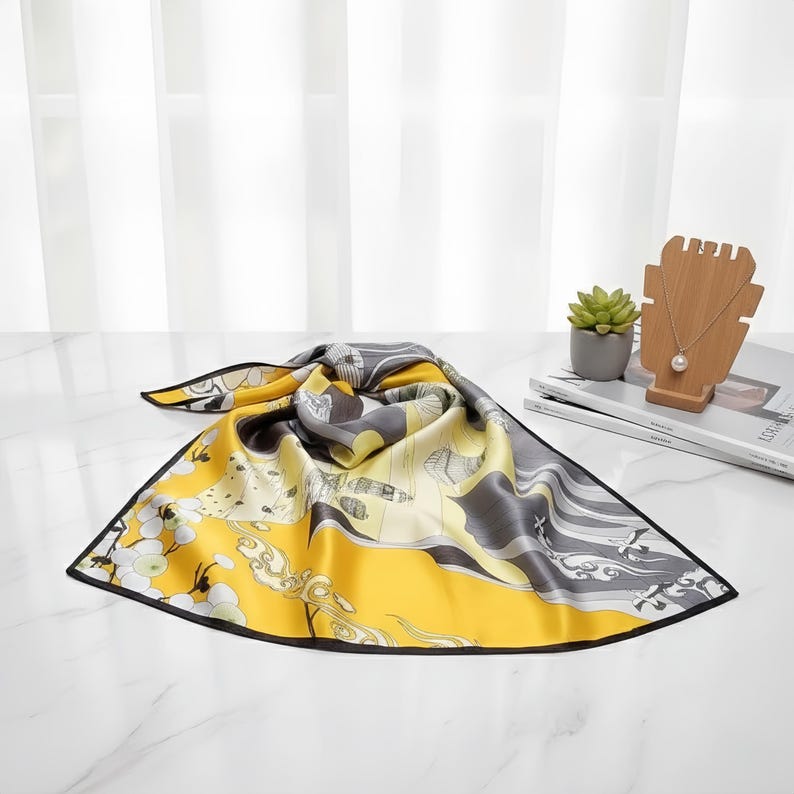 Peut inclure: Un foulard carr&eacute; en soie avec un motif floral et paysager jaune, gris et blanc. Le foulard a une bordure noire. Le foulard est pli&eacute; sur une surface blanche.