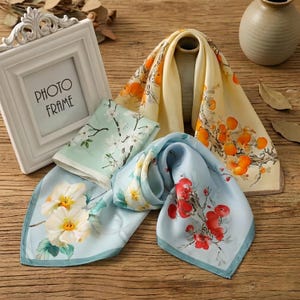 Puede incluir: Una variedad de pañuelos de seda con diseños florales y frutales. Los pañuelos presentan tonos de azul, amarillo y verde, con ilustraciones detalladas de flores y ramas. Se ve un marco de fotos blanco.