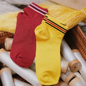 May include: A pair of ankle socks, one in deep red and one in bright yellow. Both socks feature a ribbed cuff at the top with three horizontal stripes in black, white, and red on the red sock, and black and red on the yellow sock.