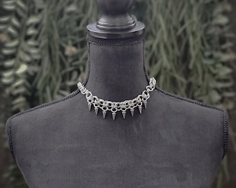 May include: A silver-colored choker necklace with a chainmail design and small, pointed spikes. The necklace is displayed on a black mannequin, with a blurred green background.