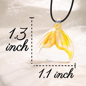 May include: A pendant necklace featuring a yellow and clear glass mermaid tail design. The pendant measures 1.3 inches long and 1.1 inches wide. The necklace has a black cord.