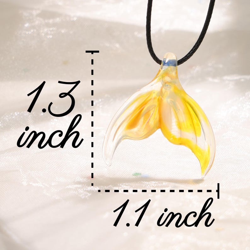 May include: A pendant necklace featuring a yellow and clear glass mermaid tail design. The pendant measures 1.3 inches long and 1.1 inches wide. The necklace has a black cord.