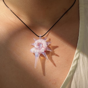 May include: A pendant necklace featuring a unique, handcrafted glass pendant. The pendant is a light pink and white spiral design with pointed edges, suspended from a black cord. The necklace is worn against a person's skin, with natural light casting a shadow.